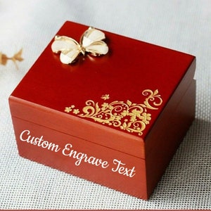 Puede incluir: Una caja de música de madera roja con un diseño de mariposa dorada y el texto "Custom Engrave Text".