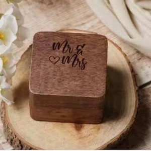 Puede incluir: Una caja de anillos de madera con las palabras "Mr & Mrs" grabadas en la parte superior. La caja est&aacute; sobre una superficie de madera.
