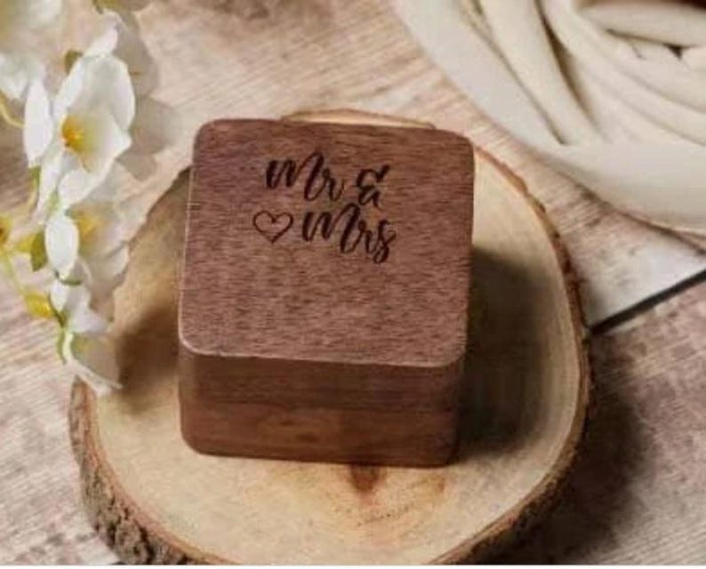 Puede incluir: Una caja de anillos de madera con las palabras "Mr & Mrs" grabadas en la parte superior. La caja est&aacute; sobre una superficie de madera.