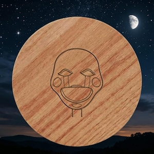 May include: A circular wooden coaster featuring a laser-engraved line art design of a stylized face with teardrop markings under the eyes, circular cheeks, and a wide, open mouth, centered on the natural light brown wood grain surface.