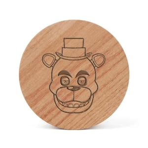 May include: A circular light brown wooden coaster featuring a laser-engraved graphic of a bear character wearing a top hat. The bear has prominent ears, a wide snout, and visible teeth. The wood grain is clearly visible across the entire surface.