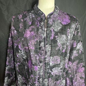 May include: A black jacket with a floral pattern in shades of purple and gray. The jacket has a stand-up collar and a full-length zipper. The fabric has a textured appearance.