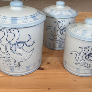 May include: A set of three off-white ceramic canisters with lids, each featuring a blue line drawing of geese wearing ribbons. The canisters have a blue plaid pattern around the top and bottom rims, with a smooth, glossy finish on the exterior.