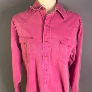 Puede incluir: Una camisa rosa brillante de manga larga con botones, dos bolsillos en el pecho y un cuello clásico. La camisa tiene detalles de estilo occidental y puños abotonados. La tela parece descolorida y desgastada.
