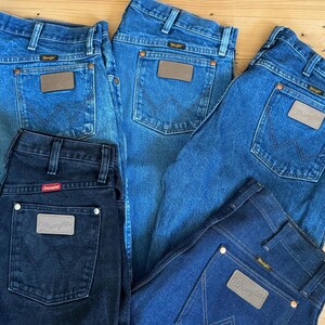 May include: A collection of Wrangler jeans in various shades of blue and black. The jeans feature classic five-pocket styling, with a leather patch on the back pocket. The jeans are arranged on a wooden surface.