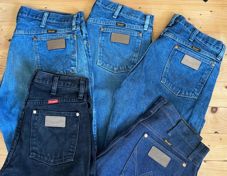 May include: A collection of Wrangler jeans in various shades of blue and black. The jeans feature classic five-pocket styling, with a leather patch on the back pocket. The jeans are arranged on a wooden surface.