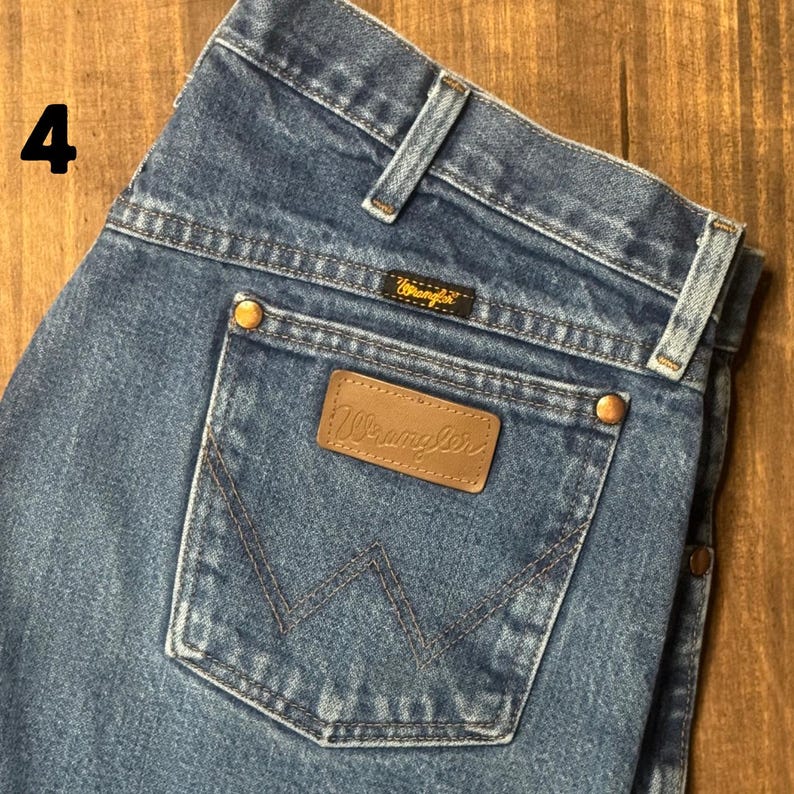 May include: Blue denim jeans with a classic five-pocket design. The back pocket features a brown leather patch with the "Wrangler" logo. The jeans have a button and zipper closure and brass rivets.
