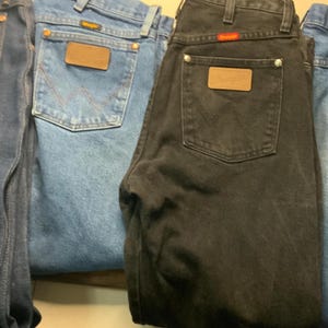 May include: Four pairs of Wrangler jeans in various shades of blue and black. Each pair features a brown leather patch on the back pocket and the Wrangler logo. The jeans are folded, showcasing the back pockets and the classic denim design.