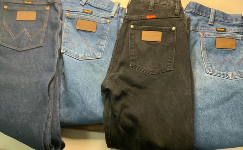 May include: Four pairs of Wrangler jeans in various shades of blue and black. Each pair features a brown leather patch on the back pocket and the Wrangler logo. The jeans are folded, showcasing the back pockets and the classic denim design.