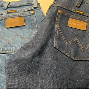 May include: Close-up of two pairs of denim jeans. One pair is a light blue wash, and the other is a dark blue wash. Both pairs feature the Wrangler logo on the back pocket and a classic five-pocket design.