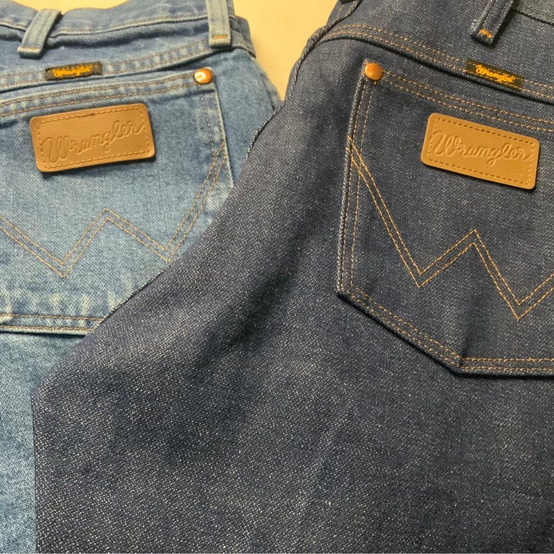 May include: Close-up of two pairs of denim jeans. One pair is a light blue wash, and the other is a dark blue wash. Both pairs feature the Wrangler logo on the back pocket and a classic five-pocket design.