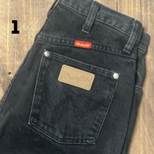 May include: Close-up of black denim jeans with a Wrangler label on the back pocket. The jeans have a red Wrangler tag above the pocket and silver rivets. The jeans are folded, showing the back of the garment.
