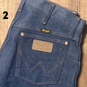 May include: Close-up of dark blue denim jeans with a Wrangler label on the back pocket. The jeans have a classic five-pocket design with copper-colored rivets and stitching. The Wrangler logo is visible on the back pocket and above the back pocket.