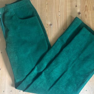 Puede incluir: Pantalones verde esmeralda con cintura alta y pernera recta. Los pantalones tienen cierre de cremallera y un único bolsillo en la parte delantera. La tela parece ser un material suave similar al ante.