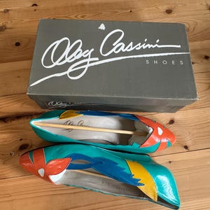 May include: A pair of teal, orange, and blue flats with a floral design, next to a gray shoe box. The box has the text "Oleg Cassini SHOES" in white script. The shoes have wooden shoe trees inside.
