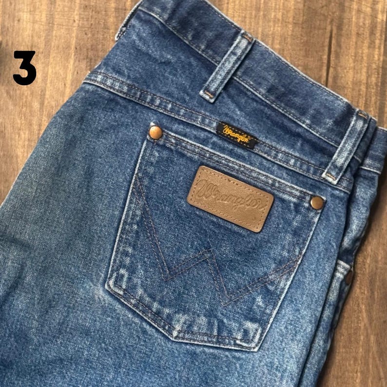 May include: Close-up of blue denim Wrangler jeans. The jeans have a brown leather patch with the Wrangler logo on the back pocket. The jeans have a classic five-pocket design and a button and zipper closure.