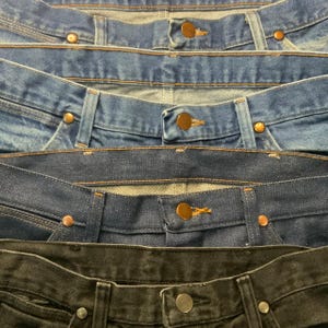 May include: Close-up of the waistbands of several pairs of denim jeans in varying shades of blue and black. The jeans have copper-colored rivets and buttons. The image shows the top portion of the jeans.