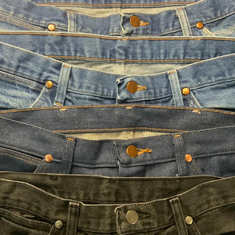 May include: Close-up of the waistbands of several pairs of denim jeans in varying shades of blue and black. The jeans have copper-colored rivets and buttons. The image shows the top portion of the jeans.