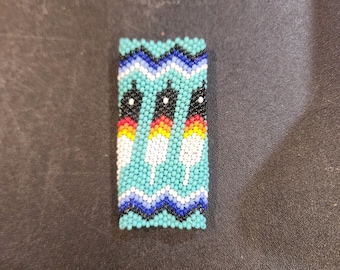 May include: A beaded rectangular ornament with a turquoise background and three black, white, yellow, and orange figures.