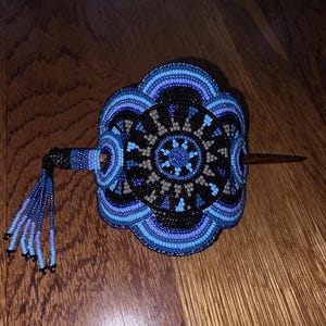 May include: A circular hair accessory featuring intricate beadwork in shades of black, light blue, and dark blue. The design includes concentric patterns and a central star-like motif. A wooden stick pierces the center, and a beaded tassel hangs from one side.