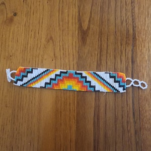 May include: A beaded bracelet with a geometric design in white, black, orange, yellow, and turquoise. The bracelet is rectangular with white loops on each end for closure. The bracelet is resting on a wooden surface.