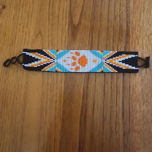 May include: Handmade beaded bracelet with a Native American design. The bracelet features a central pattern of orange and white, surrounded by turquoise, black, and white beads. The bracelet is approximately 1 inch wide and has black cord ties.