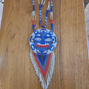 May include: A beaded necklace with a blue, white, and red face pendant. The pendant has a fringe of beads in the same colors.