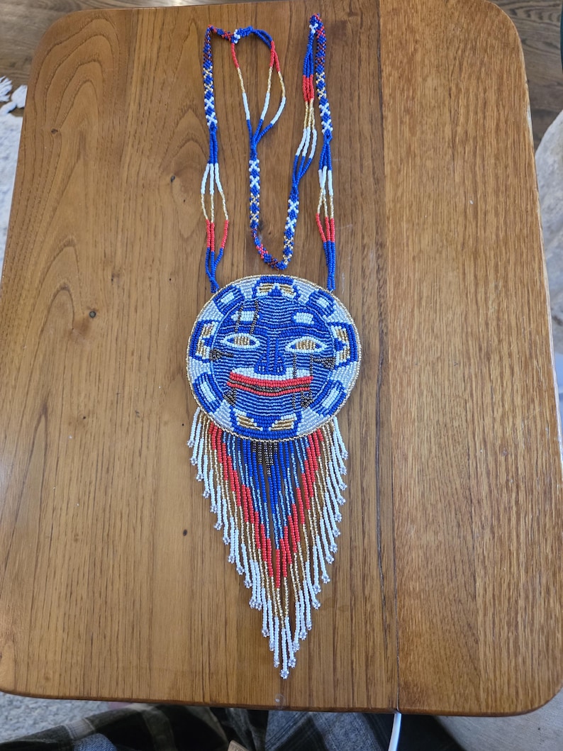 May include: A beaded necklace with a blue, white, and red face pendant. The pendant has a fringe of beads in the same colors.