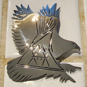 May include: A silver metal eagle sculpture with a mountain range design and a stylized mountain peak in the center. The eagle is in flight with its wings spread wide.