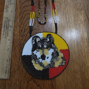May include: A circular beaded medallion necklace featuring a wolf portrait in black, white, yellow, and red beads. The pendant is approximately 3.5 inches in diameter, shown next to a ruler. The necklace includes a long beaded cord in matching colors.