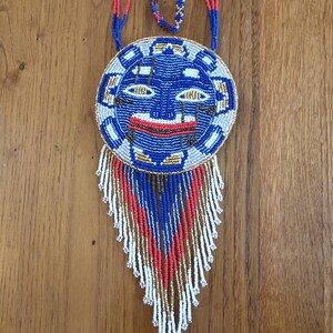 May include: A beaded necklace with a round pendant featuring a stylized face in blue, red, gold, and white. The pendant has a fringe of beads in the same colors.