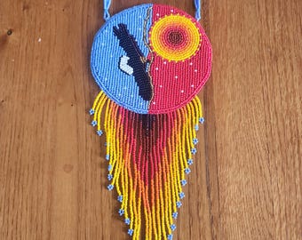 May include: A circular beaded necklace featuring a split design with a light blue side depicting a black bird silhouette and a red side with a yellow and orange sun. The piece includes long, multi-colored fringe strands in yellow, orange, and red.