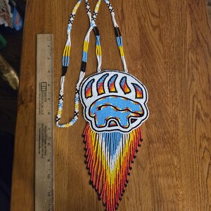 May include: A beaded necklace featuring a bear paw pendant with a blue bear silhouette, accented by red, orange, yellow, and white fringe. The necklace includes a long beaded strap and is shown next to a 12-inch ruler for scale reference.