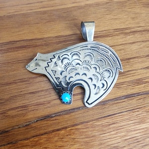 May include: Silver-toned bear pendant with intricate etched details and a turquoise stone eye. The pendant has a loop for a chain and is displayed on a wooden surface. The bear design is a popular motif in Native American jewelry.