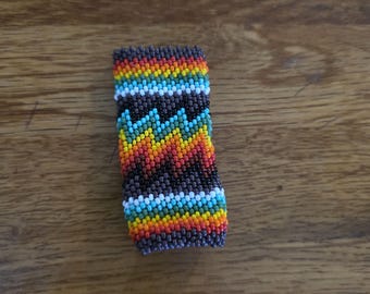May include: A rectangular beaded bracelet with a vibrant pattern of rainbow colors, including red, orange, yellow, green, blue, and white. The bracelet has a dark brown background and a zig-zag design in the center.