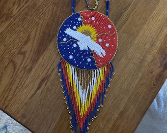 May include: A beaded necklace with a large circular pendant featuring a white eagle design against a blue and red background. The necklace has blue, red, yellow, and gold beads, with a fringe of beads at the bottom.