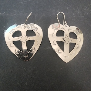 May include: A pair of silver heart-shaped earrings. Each earring features a cut-out cross design with a star in the center. The edges are decorated with small, curved markings. The earrings have a hook closure.