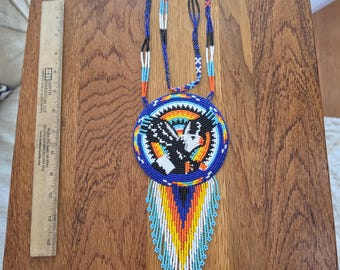 May include: A beaded medallion necklace featuring a central eagle design in black, white, and orange, surrounded by blue, yellow, and red geometric patterns. The necklace includes a long, multi-colored fringe and a 12-inch ruler for scale.