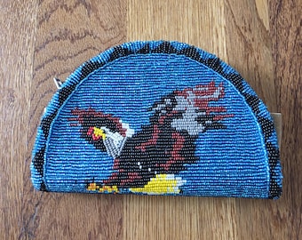 May include: A semi-circular coin purse featuring a beaded design of a bald eagle in flight. The eagle is rendered in black, white, red, and yellow beads against a bright blue background, with a black beaded border along the curved edge.