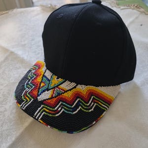 May include: A black baseball cap with a beaded brim. The brim features a colorful geometric design with red, orange, yellow, green, blue, and white beads. The cap is made of a dark fabric.