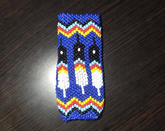 May include: A rectangular beaded item with a blue background and a zigzag pattern at the top. The design features three white feather-like shapes with black and white accents. The item is likely a small pouch or case.