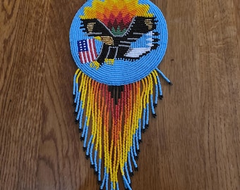 May include: Handmade beaded necklace featuring a central round pendant with a blue background. The pendant showcases an eagle design with the American flag. The necklace has a fringe of yellow, orange, and blue beads, and a beaded chain.