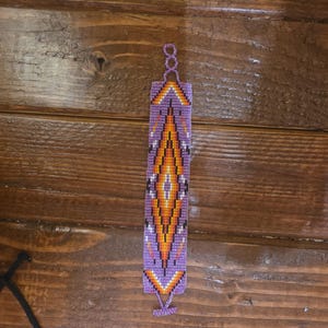 May include: A beaded bracelet with a geometric design in shades of purple, orange, yellow, and white. The bracelet features a central diamond pattern and a loop closure. The background is a wooden surface.