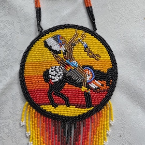 May include: A beaded necklace with a circular pendant featuring a Native American rider on horseback. The pendant has a yellow, orange, and black color scheme, with a black border and beaded fringe.