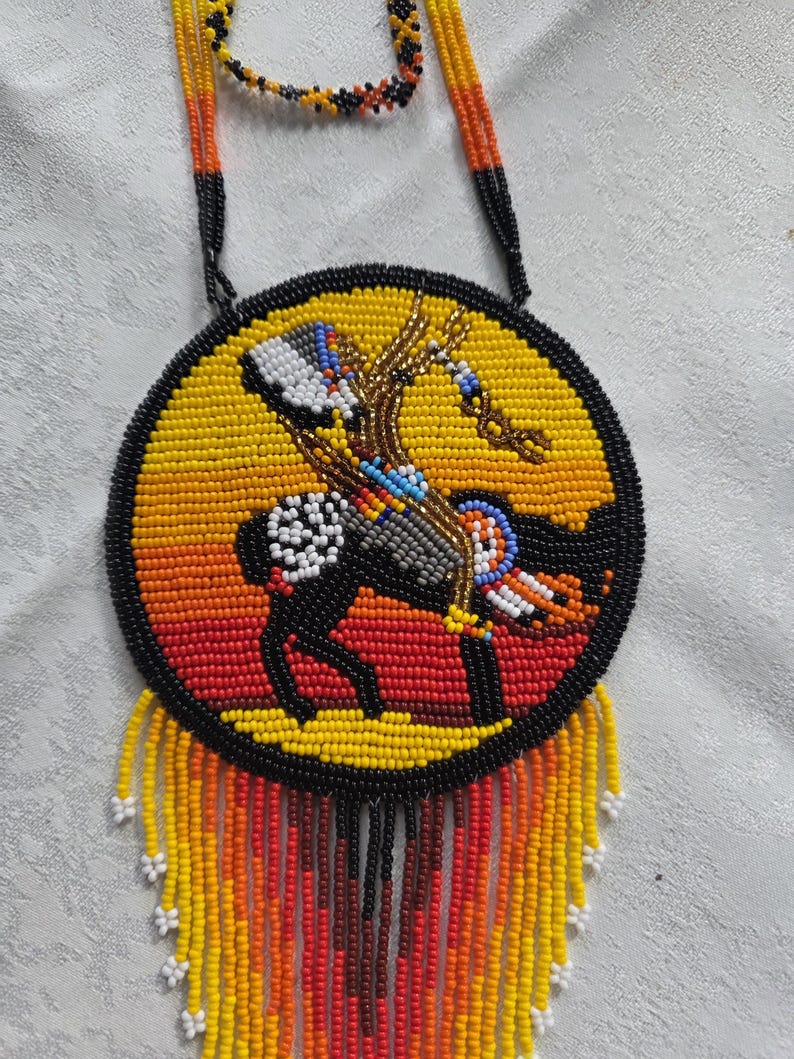 May include: A beaded necklace with a circular pendant featuring a Native American rider on horseback. The pendant has a yellow, orange, and black color scheme, with a black border and beaded fringe.