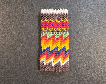 May include: Rectangular beaded item with a white top and a brown base. The design features zig-zag patterns in red, yellow, blue, green, and orange. The beads are small and tightly packed, creating a textured surface.