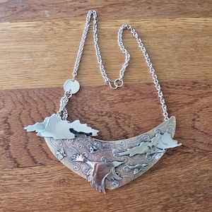 May include: A crescent-shaped metal necklace featuring a textured bronze-toned background with engraved swirling patterns. The piece includes polished silver-toned metal overlays depicting clouds and birds in flight, suspended from a silver-toned chain.