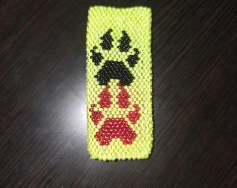 May include: A rectangular, beaded item in a bright yellow-green color. The item features two paw prints, one black and one red, made of small beads. The paw prints are centered on the item.