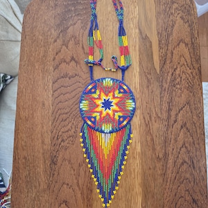 May include: A handcrafted beaded necklace featuring a large circular pendant with a vibrant starburst pattern in blue, red, yellow, and green. The pendant is accented with long, multi-colored beaded fringe strands that taper to a point.