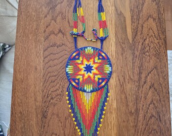 May include: A handcrafted beaded necklace featuring a large circular pendant with a vibrant starburst pattern in blue, red, yellow, and green. The pendant is accented with long, multi-colored beaded fringe strands that taper to a point.